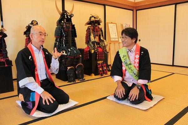 鎧兜の前で黒い法被を着た原田伸郎さんが担当者から話を聞いている写真